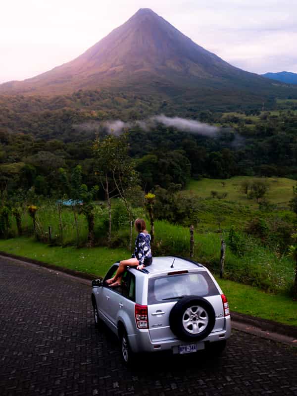Lady sitting over a car enjoying the sunset while sun goes behind the Arenal Volcano by Costa Rica Unexplored.